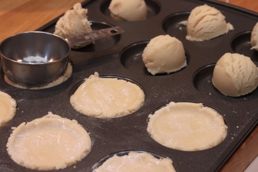sugar cookie dough being pressed into wells of a deep dish cookie sheet with a measuring cup