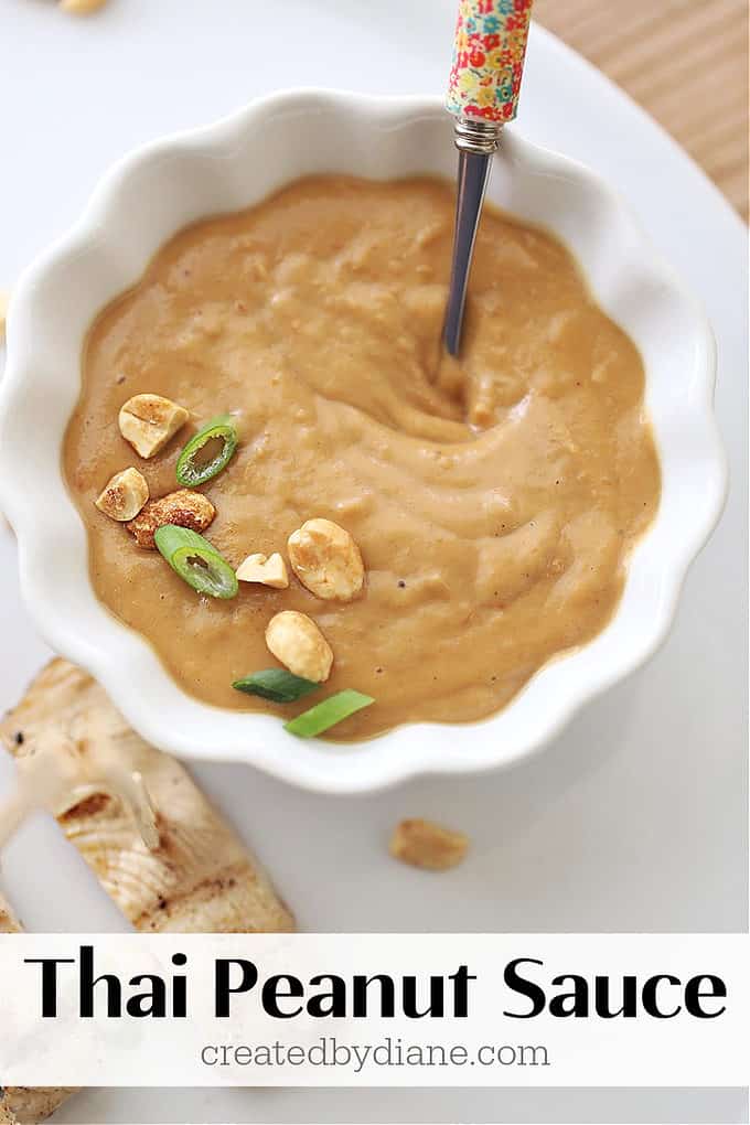 White bowl with wavy edge filled with thick thai peanut sauce topped with scallions and chopped peanuts and a small spoon on a white plate with grilled chicken pieces