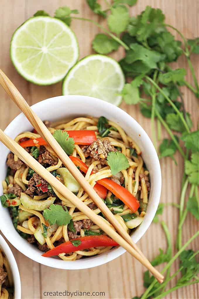 white bowl with chopsticks, ground beef, spaghetti, red pepper, bok choy, sliced limes and cilantro
