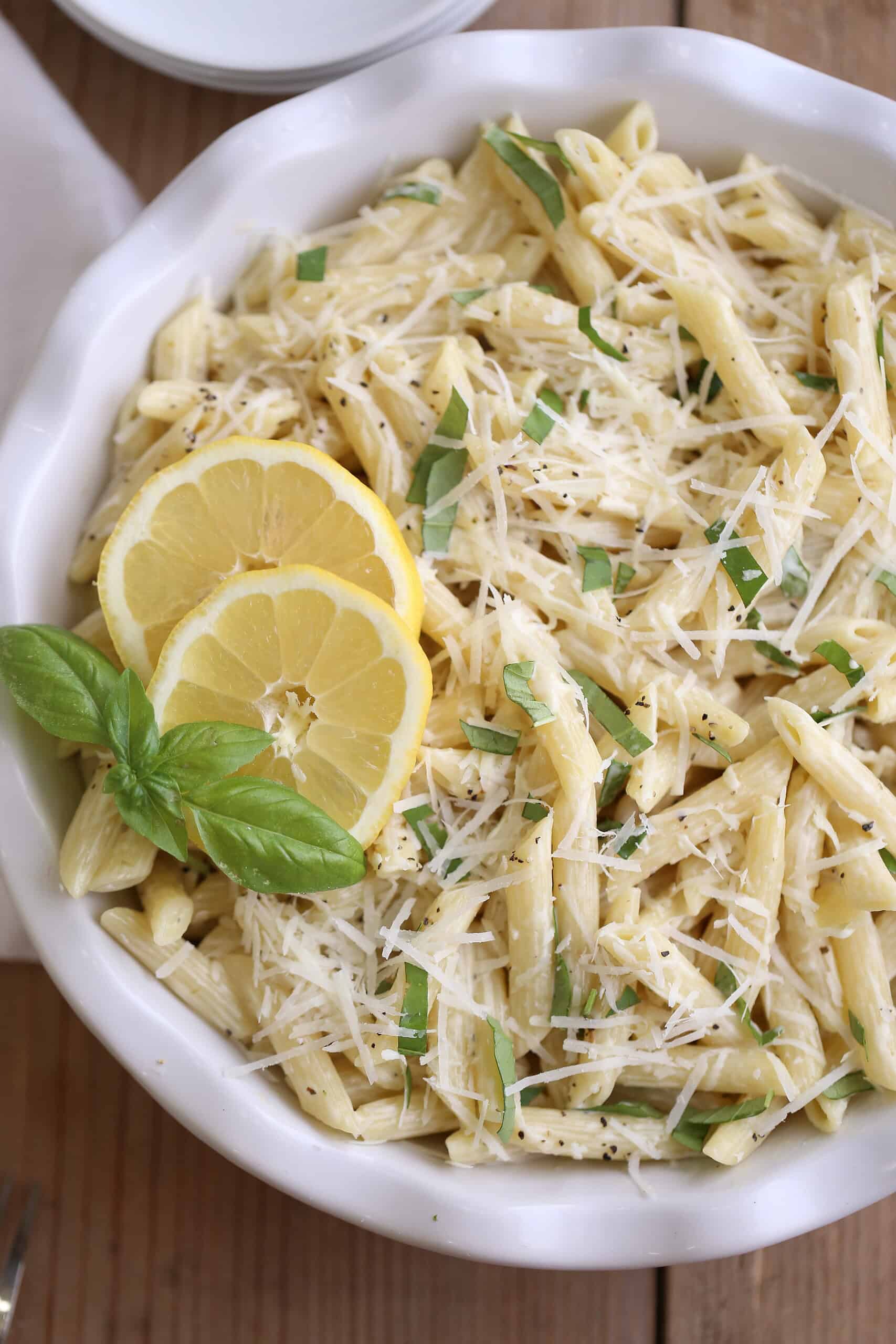 White bowl, fluted edge filled with a creamy lemon pasta salad, with lemon garnish and basil, topped with shredded Romano cheese